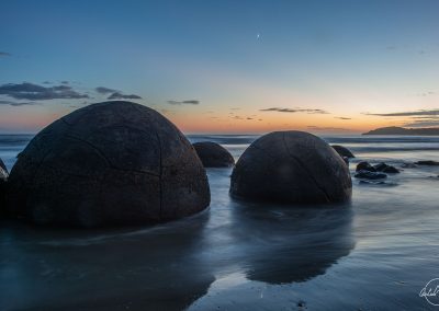 Two spherical stones on the beach in Moeraki at sunrise with the moon in the sky