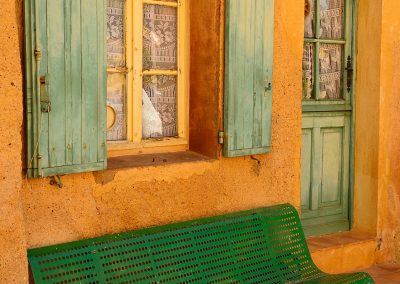 Green wooden bench in front of a yellow house with light green shutters in Roussillon