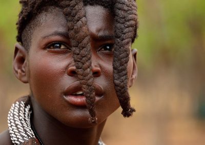 Portrait of a teenage himba with braids on her front and green trees in background