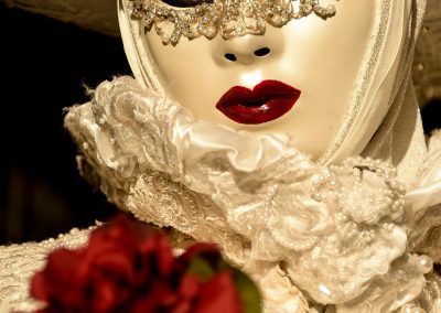 Woman in a white Venitian mask and costume with red lips, green eyes and a red rose in the foreground