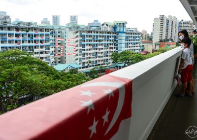 Mother and child wearing in mask and waiting for mobile columns from their HDB balcony with Singapore flag lying in foreground and 55 building in background in Toa Payoh